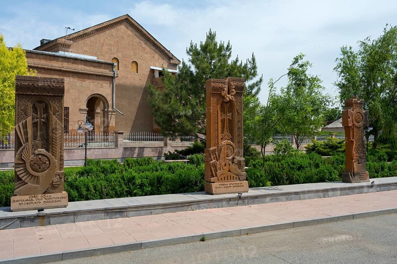 Three ornately decorated khachkars in front of a historic building, surrounded by green trees and well-tended hedges, Khachkar, Cross Stone, Aghavnatun, Armavir Province, Armenia