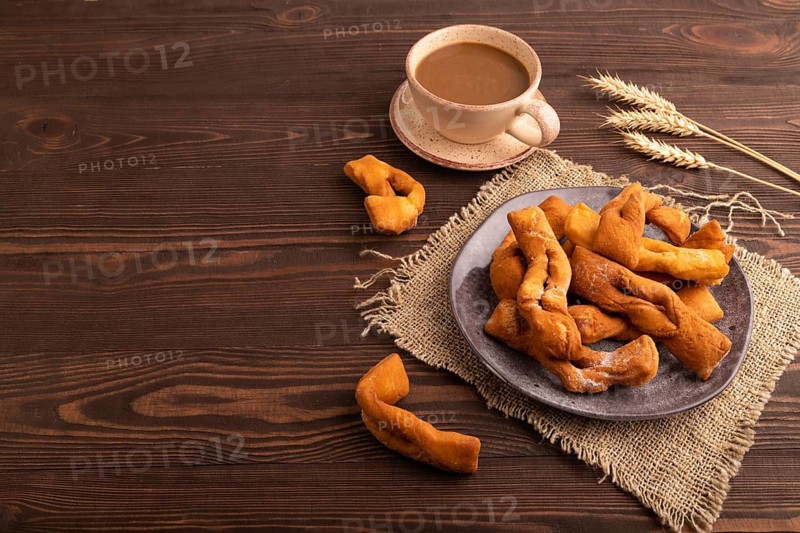 Crunchy biscuit Brushwood cookies sprinkled with powdered sugar on brown wooden background and linen textile, cup of coffee, side view, copy space