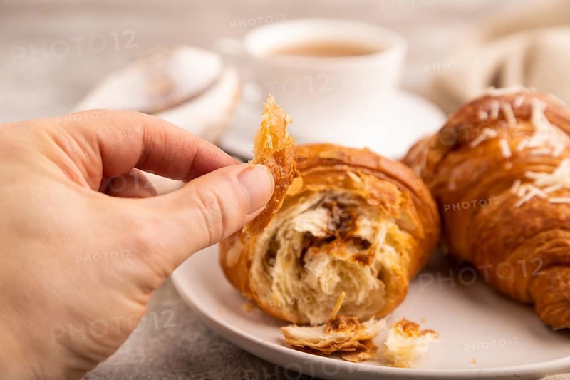 Croissant on white plate with hand on brown concrete background and linen textile, cup of coffee, side view, close up, selective focus
