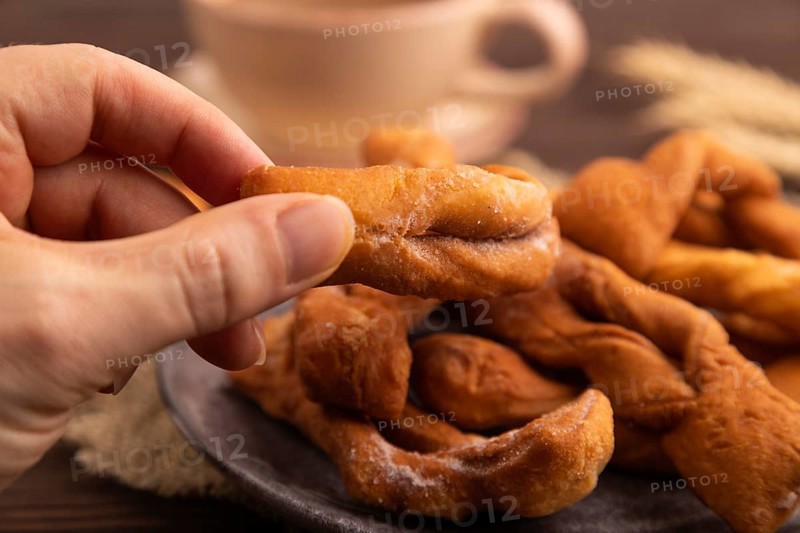 Crunchy biscuit Brushwood cookies with hand sprinkled with powdered sugar on brown wooden background and linen textile, cup of coffee, side view, close up, selective focus