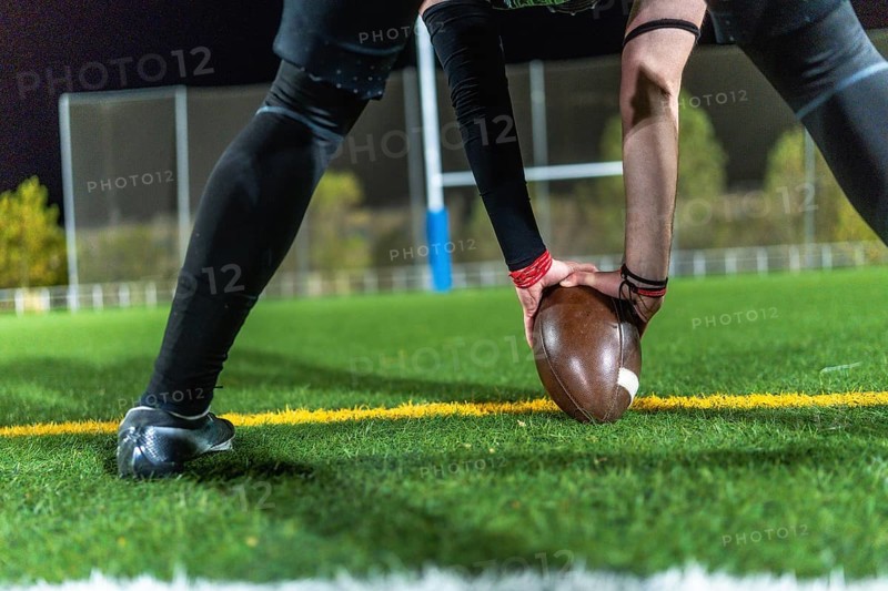 American football player in dark uniform with arm sleeves placing a brown ball on green turf at night, low angle close up capturing focus before kickoff