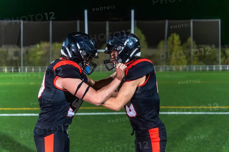 Two american football players in black and orange uniforms collide in a gritty close up on green turf under stadium lights, locked in a physical, high energy struggle