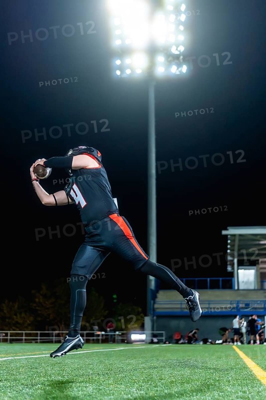 American football player in black and orange uniform catching an airborne football on a green turf field at night, illuminated by bright stadium lights during an evening game