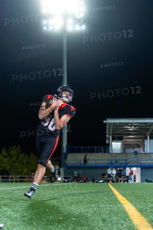 American football player in uniform and helmet catching the football on a green turf field at night under bright stadium lights, with blurred spectators in the stands