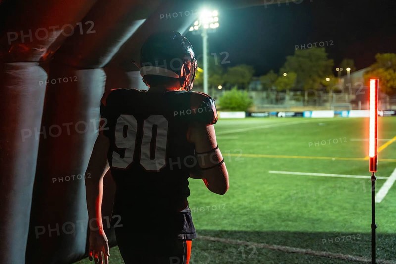 American football player in jersey number 90 emerges from an inflatable tunnel onto a floodlit stadium field at night, low angle back view capturing power and anticipation