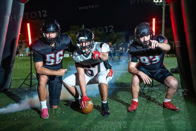 Three determined american football players, one holding the ball, emerging from an inflatable tunnel onto a stadium field at night, ready for game action