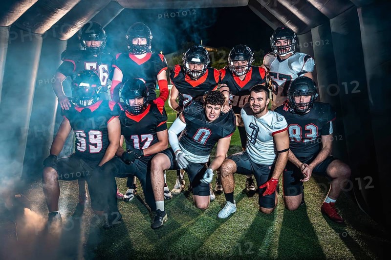 American football team poses united in helmets and pads, emerging from a smoky stadium tunnel under night lights, focused and ready for competition on the turf