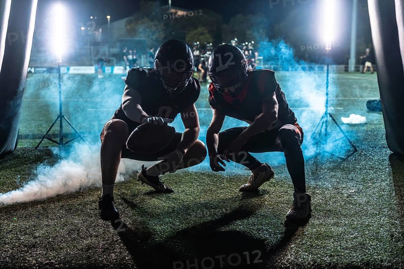 Two american football players in full gear, crouching and ready for action, emerging from a dark tunnel onto a stadium field at night with dramatic smoke and bright lights