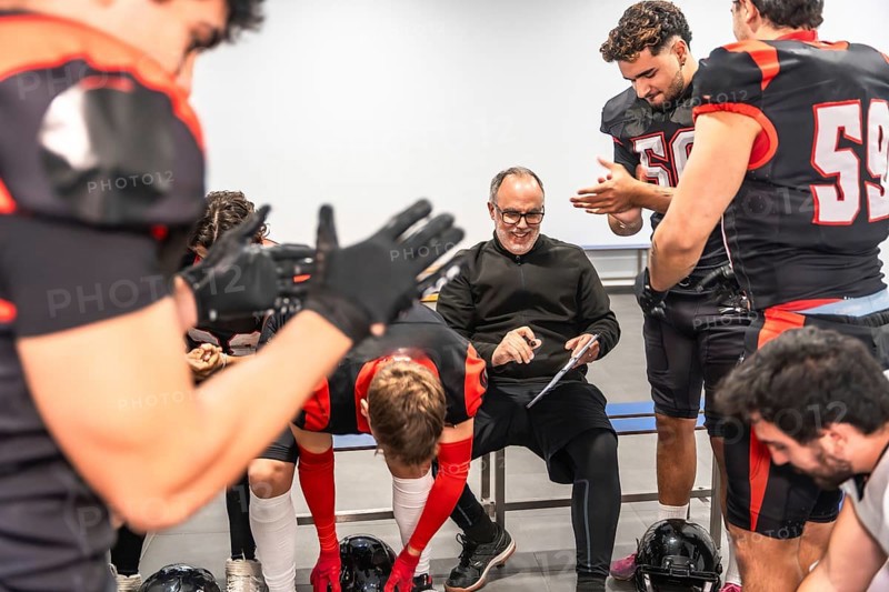 American football coach smiling, discussing strategy, and preparing for a game with diverse male players dressed in uniforms and protective gear inside a locker room