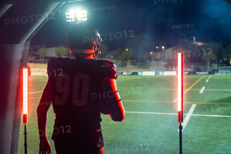 American football player in full uniform and helmet emerging from a tunnel onto a floodlit stadium field at night, holding the ball and poised to charge into play