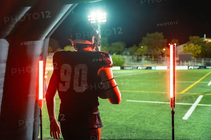 American football player wearing helmet and uniform with number 90, holding a football, walking through a tunnel onto a floodlit stadium field at night for a game
