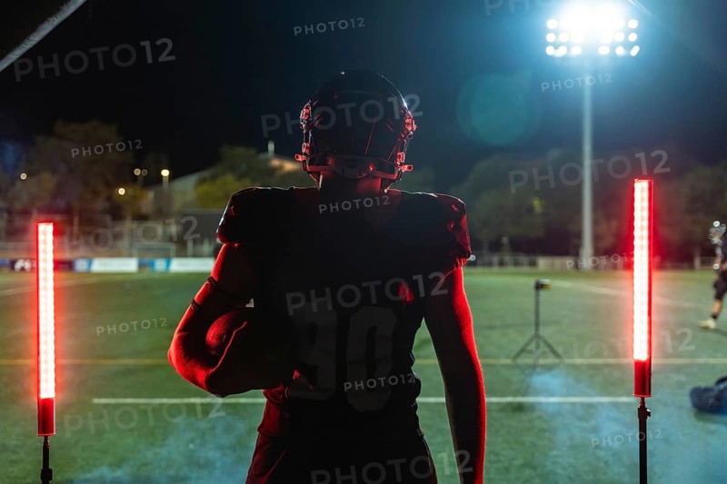 Football player stands on stadium field at night, helmet and uniform on, holding ball and facing bright red glowing lights, poised and ready for intense competition and victory