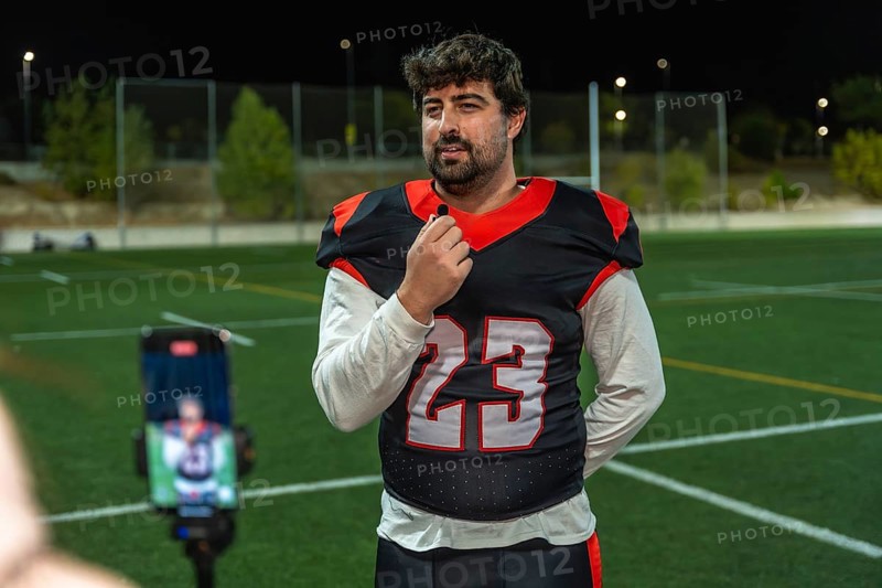 American football player wearing a jersey with number 23 giving an interview on a sports field at night while speaking into a lapel microphone, with a smartphone recording the content on a tripod