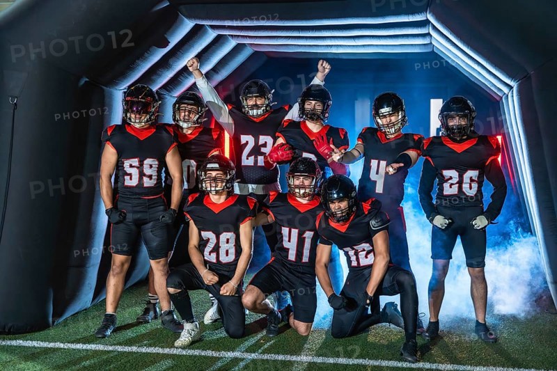 American football team players standing ready to enter the stadium through a tunnel, wearing helmets and black and red uniforms, posing with excitement before the game
