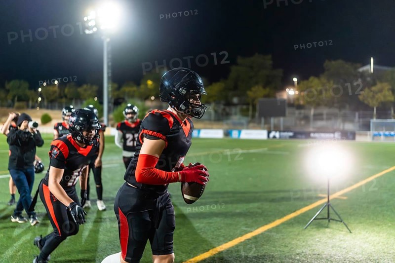Female american football player training on a stadium field at night, wearing a helmet and uniform, holding the football and running, concentrating on the game