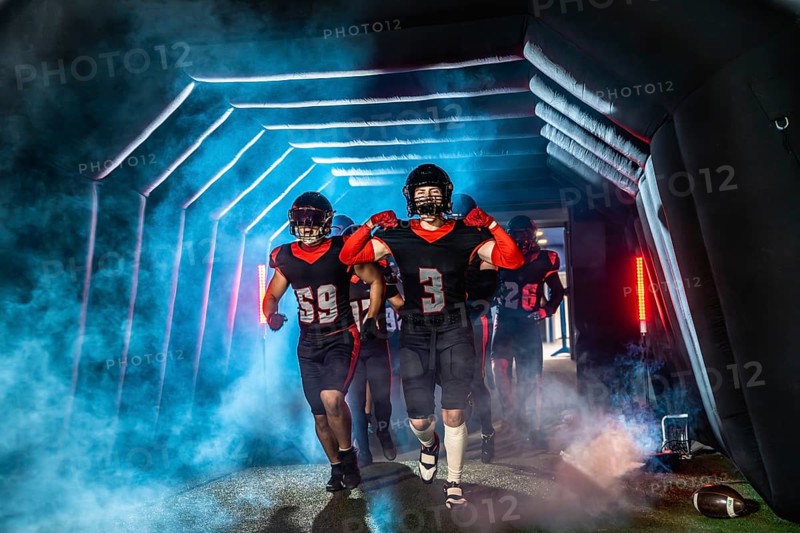 American football players in black and red uniforms and helmets running through a dramatic, smoke filled stadium tunnel with glowing lights, preparing for a game