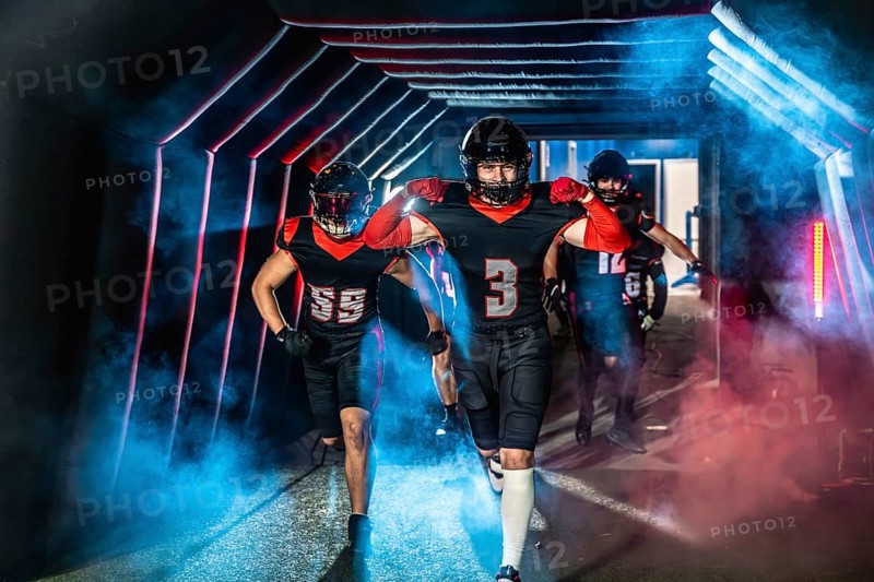 American football players are running forward from a dark stadium tunnel filled with blue and red smoke, showing determination and readiness for competition on the field