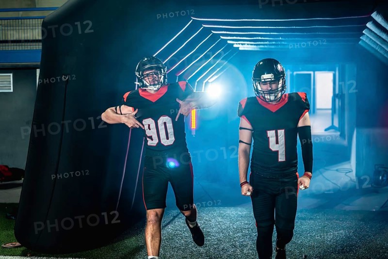 American football players in black and red uniforms and helmets are making an entrance, emerging from a dark tunnel illuminated with blue light, ready for a game on the field below