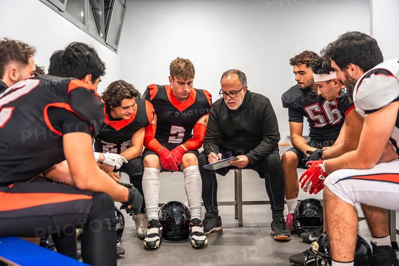 American football coach discussing strategy with his team of male players in the changing room, analyzing tactics and giving instructions before a game