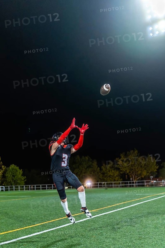 American football player in uniform and helmet is jumping to catch a football during a night game on a sports field under bright stadium lights, demonstrating athletic skill