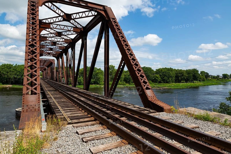 Railroad tracks leading to old rusted Terrebonne Pratt truss railroad bridge over Des Mille-Iles river in summer, Laval, Quebec, Canada