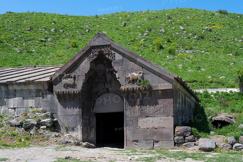 Old stone building with detailed decorations in front of a lush green hill on a sunny day, Orbelian's Caravanserai, Selim Caravanserai, located at Vardenyats Pass or Selim Pass, Vayots Dzor Province, Armenia