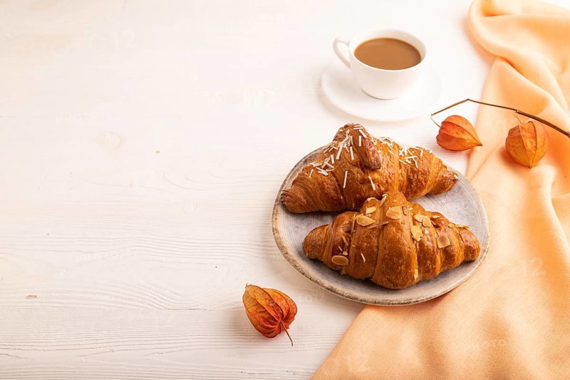 Croissant on blue plate on white wooden background and orange linen textile, cup of coffee, side view, copy space