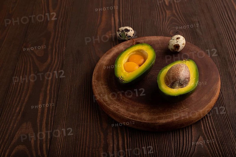 Two Avocado halves with pit and Egg yolk on round wooden cutting board on brown wooden background, side view, copy space, minimalism, low key