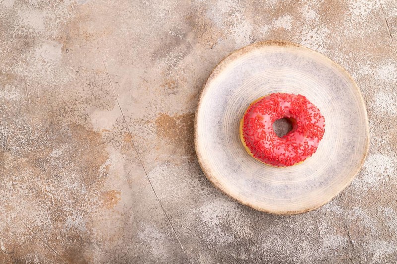 Pink Donut with sprinkles on blue ceramic plate on brown concrete background, top view, flat lay, copy space, minimalism