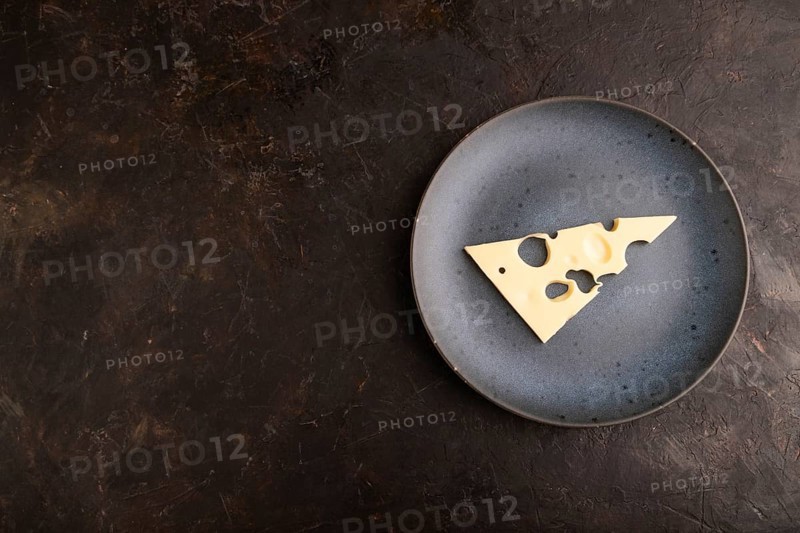 Piece of cheese with holes on blue plate on black concrete background, top view, flat lay, copy space, minimalism