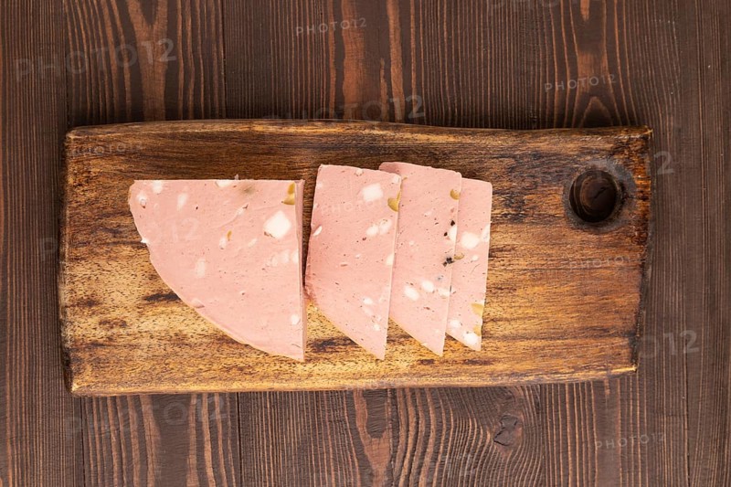 Mortadella sausage on wooden cutting board with pepper and herbs on brown wooden background. Top view, flat lay, close up