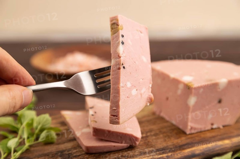 Mortadella sausage with hand on wooden cutting board with pepper and herbs on brown wooden background. Side view, close up, selective focus