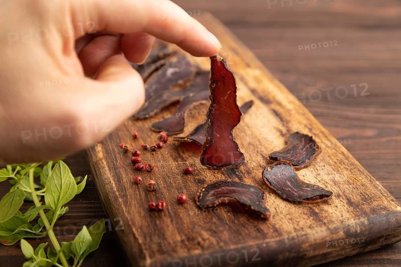 Armenian Basturma dried meat with hand on wooden cutting board with pepper and herbs on brown wooden background. Side view, close up, selective focus