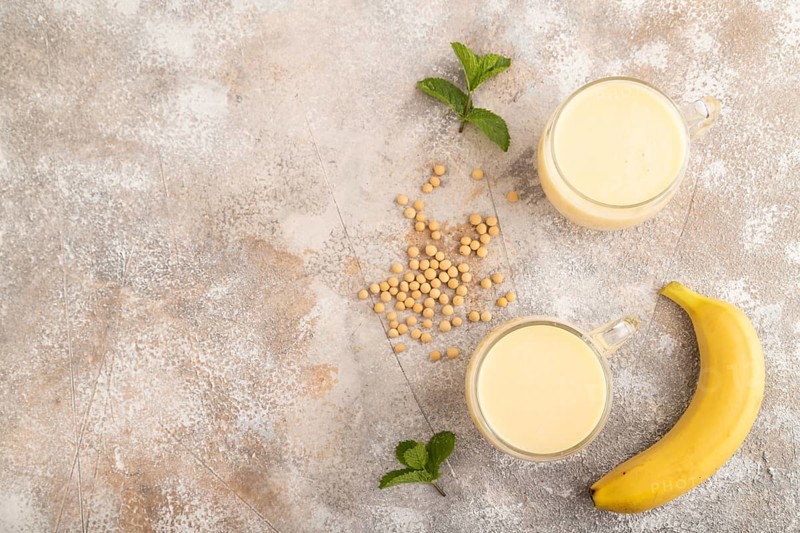 Organic non dairy banana and soy milk in glass on brown concrete background. Vegan healthy food concept, flat lay, top view, copy space