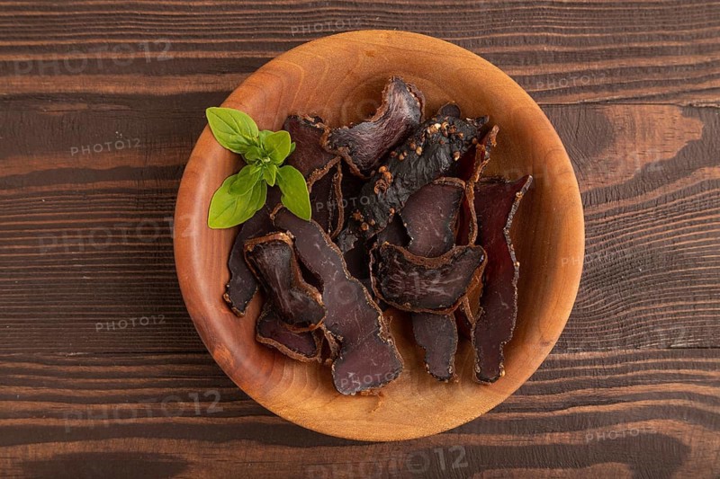 Armenian Basturma dried meat on wooden bowl with pepper and herbs on brown wooden background. Top view, flat lay, copy space