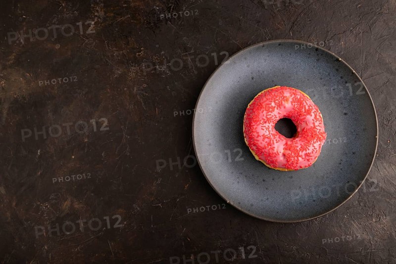 Pink Donut with sprinkles on blue ceramic plate on black concrete background, top view, flat lay, copy space, minimalism
