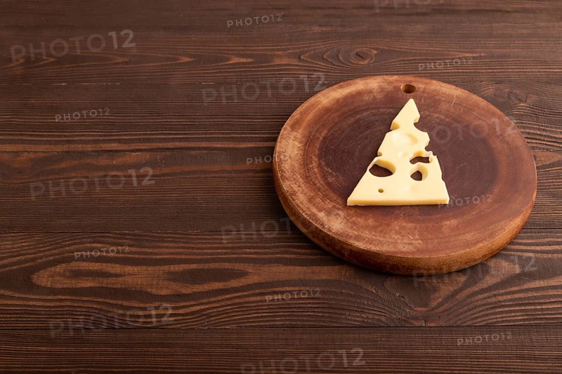 Piece of cheese with holes on cutting board on brown wooden background, side view, copy space, minimalism