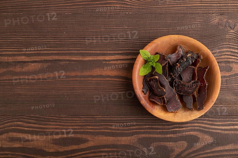 Armenian Basturma dried meat on wooden bowl with pepper and herbs on brown wooden background. Top view, flat lay, copy space