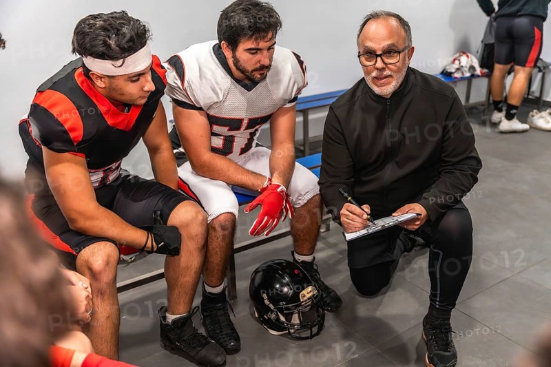 American football coach on a knee formulating game strategy and providing guidance on a clipboard to two players sitting on a locker room bench, creating a scene of teamwork and preparation