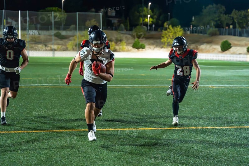American football players competing on a lit field at night, a player carrying the ball and sprinting to gain yards while opponents are pursuing him during intense action