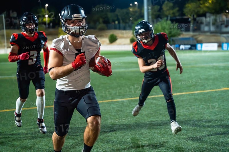 American football players, wearing protective helmets and pads, are running on a green turf field under bright stadium lights during an intense training session or evening game