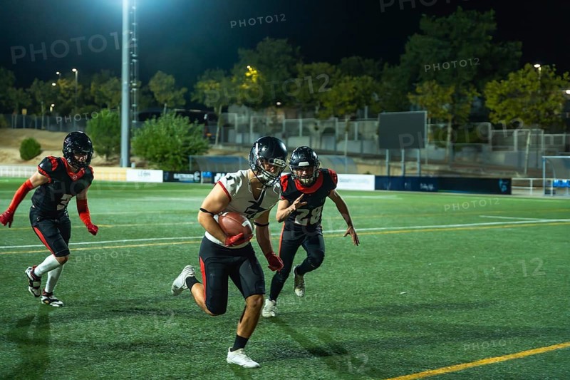 American football player vigorously running with the ball on a green field during a night game, two opposing players chasing in pursuit, highlighting action and competitive athletic performance