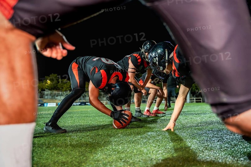 American football players line up on the field at night, a center snapping the ball, preparing for an intense moment of competitive sports action during a game