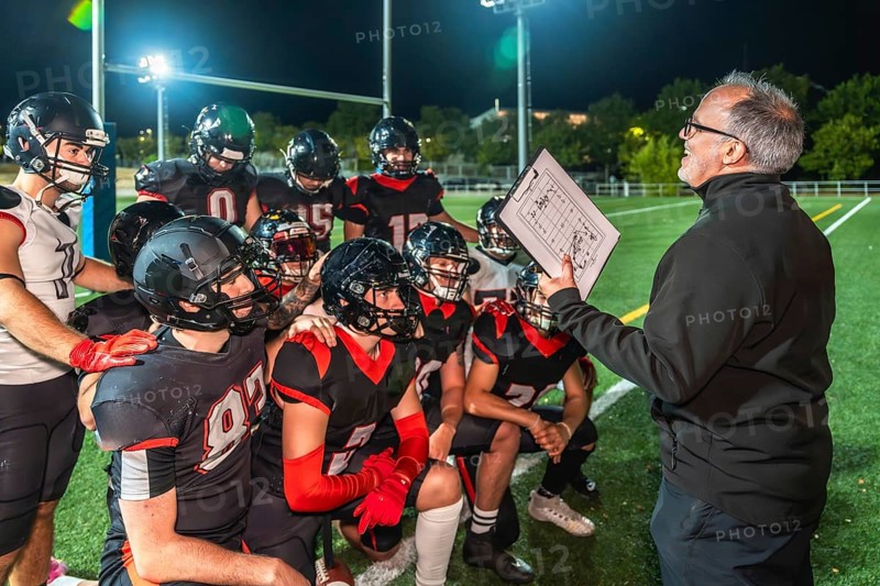 American football coach holds clipboard with play diagram, instructing young athletes in full uniform and helmets during a nighttime huddle on bright stadium turf field