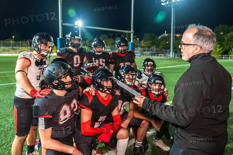 American football coach discussing strategy with his team of players in uniform and helmets during a nighttime practice or game on a green turf field under bright stadium lights