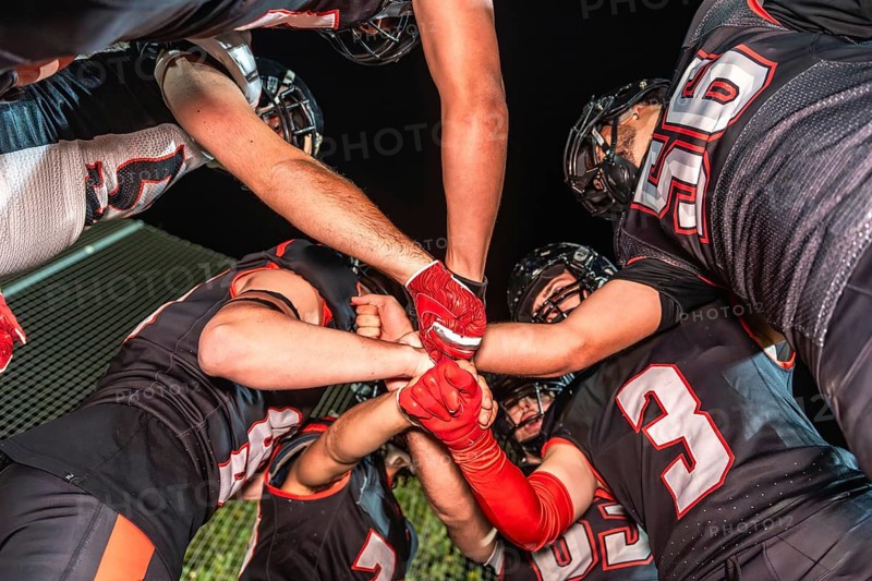 Football teammates in a night huddle, gloved fists stacked in the center, conveying unity, teamwork, power and focused strategy before the big play under stadium lights