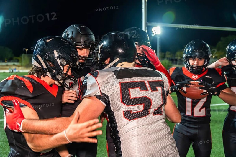 American football players in black and red uniforms with helmets and shoulder pads huddling on a green turf field during a night game, discussing strategy and demonstrating teamwork