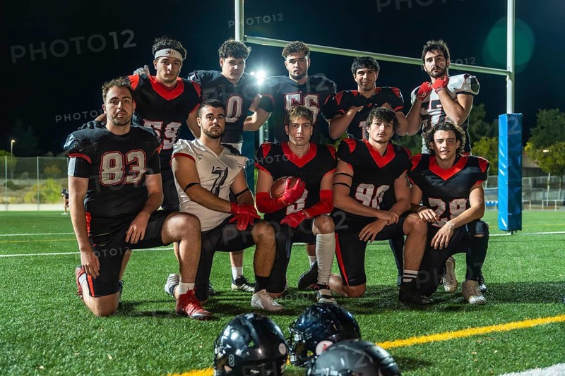 American football team in uniforms posing on illuminated turf at night, holding a ball and celebrating teamwork, focus and athletic determination before the game