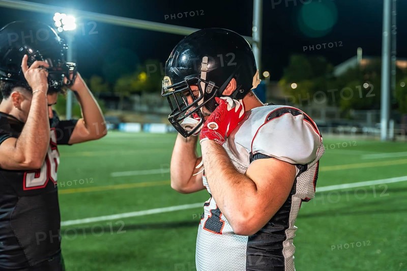 American football players adjusting their helmets on illuminated green field, preparing for intense training session or competitive game under the night sky