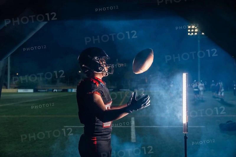 American football player in helmet and uniform lunging to catch a pass under stadium lights, smoke drifting across the night field in an intense, action packed moment